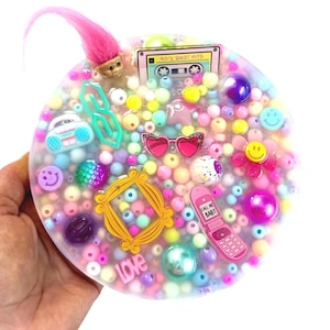 Op de afbeelding: Een ronde, heldere hars coaster gevuld met kleurrijke kralen en 90's thema bedels. Inclusief een roze trollenpop, een cassettebandje, hartvormige zonnebril, een roze klaptelefoon en een gouden fotolijst. De coaster heeft het woord "LOVE" erop.