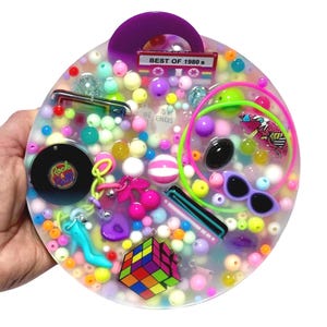 May include: A round, clear resin tray filled with colorful beads and 1980s-themed charms. Charms include a Rubik's Cube, sunglasses, a cassette tape that says "Best of 1980s", and a "Rock & Roll" record. The tray has a purple handle.
