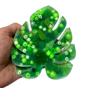 Puede incluir: Un objeto decorativo en forma de hoja de resina verde, lleno de cuentas verdes de varios tonos y pequeñas inclusiones de hojas. La resina translúcida permite ver las cuentas. El artículo se sostiene en una mano sobre un fondo blanco.