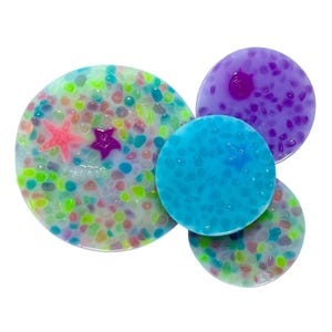 May include: A collection of colorful, round resin art pieces. The largest piece is a light green with a variety of small, colorful stones and two star-shaped accents. Other pieces are blue, purple, and another green, all with unique textures.