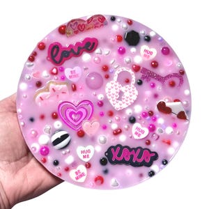 Puede incluir: Un posavasos de resina rosa con elementos decorativos incrustados. El posavasos presenta la palabra "love", corazones y las frases "Be Mine", "Hug Me" y "XOXO". Está decorado con cuentas y otros pequeños adornos.