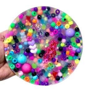 May include: A round, clear resin coaster filled with colorful beads, including pink, purple, green, and blue. The coaster also features small teddy bear and heart-shaped charms, creating a playful and vibrant design.