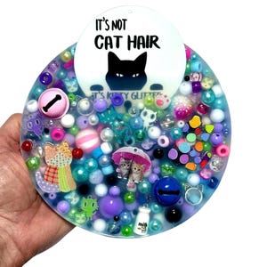 Puede incluir: Una pieza de arte redonda de resina transparente llena de cuentas, abalorios y purpurina de colores. La pieza presenta una silueta de gato negro y el texto "It's Not Cat Hair". Varios adornos con temática de gatos están dispersos.