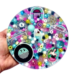 Puede incluir: Un posavasos redondo de resina con temática de los años 50, con una gramola, un letrero de restaurante y un coche clásico. El posavasos está lleno de cuentas y abalorios de colores en tonos rosa, azul y amarillo. También incluye un disco de vinilo en blanco y negro.