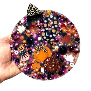 Puede incluir: Un posavasos redondo de resina transparente con temática de Halloween. El posavasos está lleno de varias cuentas en tonos morados, naranjas y dorados. Los elementos decorativos incluyen calabazas, hojas y un lazo con estampado de leopardo. Las palabras "IT'S FALL Y'ALL" son visibles.