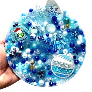 Puede incluir: Adorno navideño redondo, azul y blanco, con varios elementos decorativos. Presenta un pingüino con un suéter, copos de nieve y un regalo. El adorno está lleno de cuentas azules y blancas y tiene un acabado brillante.
