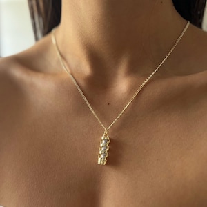 May include: A gold-colored necklace with a cylindrical pendant. The pendant features a series of gold and silver-colored rectangular bars. The necklace is worn around the neck.