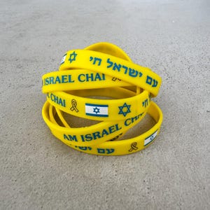 Puede incluir: Un conjunto de pulseras de goma amarillas con las palabras "Am Israel Chai" y "Chai" en hebreo e inglés, junto con una estrella de David y una cinta amarilla.