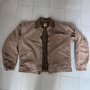 Op de afbeelding: Een bruine werkjas met een corduroy kraag en een geruite voering. De jas heeft twee voorzakken en een kleine vierkante patch op de borst. De jas is gemaakt van een duurzaam, versleten materiaal.