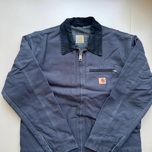 Könnte beinhalten: Marineblaue Arbeitsjacke mit schwarzem Kragen und durchgehendem Reißverschluss. Die Jacke hat eine kleine Reißverschlusstasche auf der Brust und ein quadratisches Logo-Patch. Die Jacke ist aus strapazierfähigem Material gefertigt und hat einen klassischen Arbeitsstil.