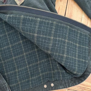 May include: A dark green denim jacket with a plaid lining. The jacket has a zipper closure and two buttoned pockets. The Carhartt logo is visible on the left chest pocket.