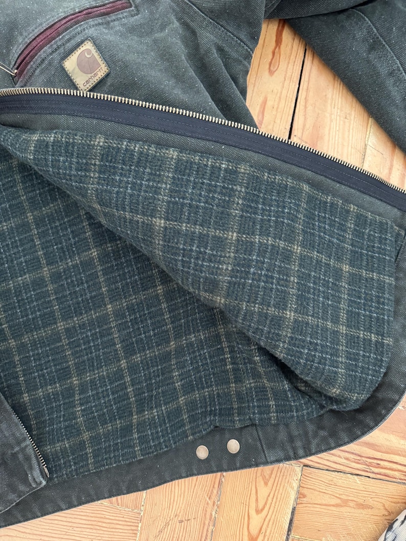 May include: A dark green denim jacket with a plaid lining. The jacket has a zipper closure and two buttoned pockets. The Carhartt logo is visible on the left chest pocket.