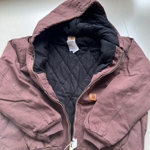 May include: A mauve-colored hooded jacket with a black quilted interior lining. The jacket features a front metal zipper closure, ribbed cuffs and hem, and a small rectangular Carhartt brand patch on the left chest area.