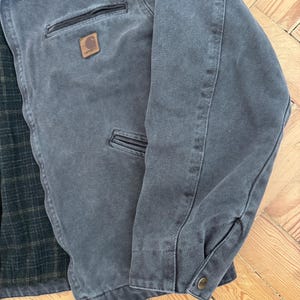 May include: A gray Carhartt jacket with a brown leather patch on the chest. The jacket has a dark gray collar, two front pockets, and a buttoned cuff. The interior lining is a green and black plaid pattern.