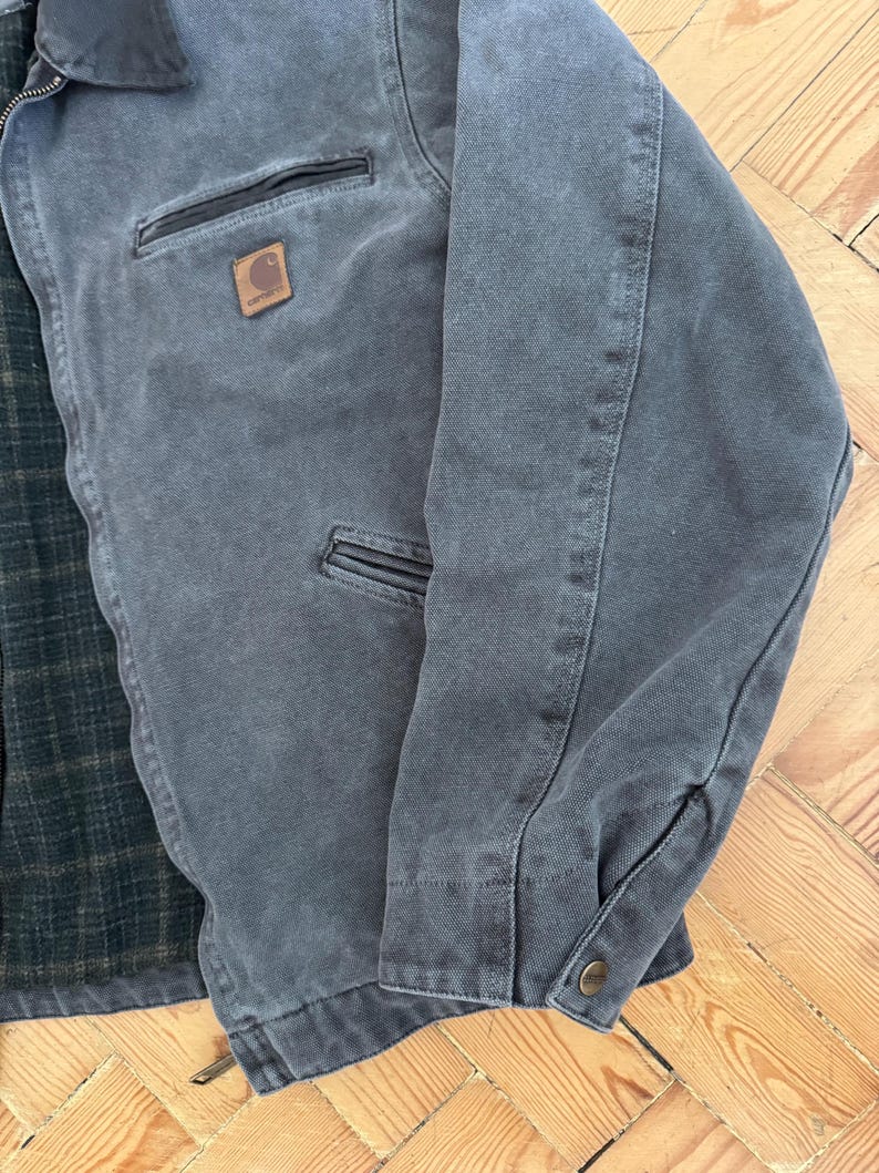 May include: A gray Carhartt jacket with a brown leather patch on the chest. The jacket has a dark gray collar, two front pockets, and a buttoned cuff. The interior lining is a green and black plaid pattern.