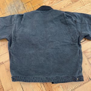 May include: A dark gray work jacket with a black collar. The jacket has a button closure and two front pockets. The fabric appears to be a durable cotton or canvas material, suitable for work or casual wear.