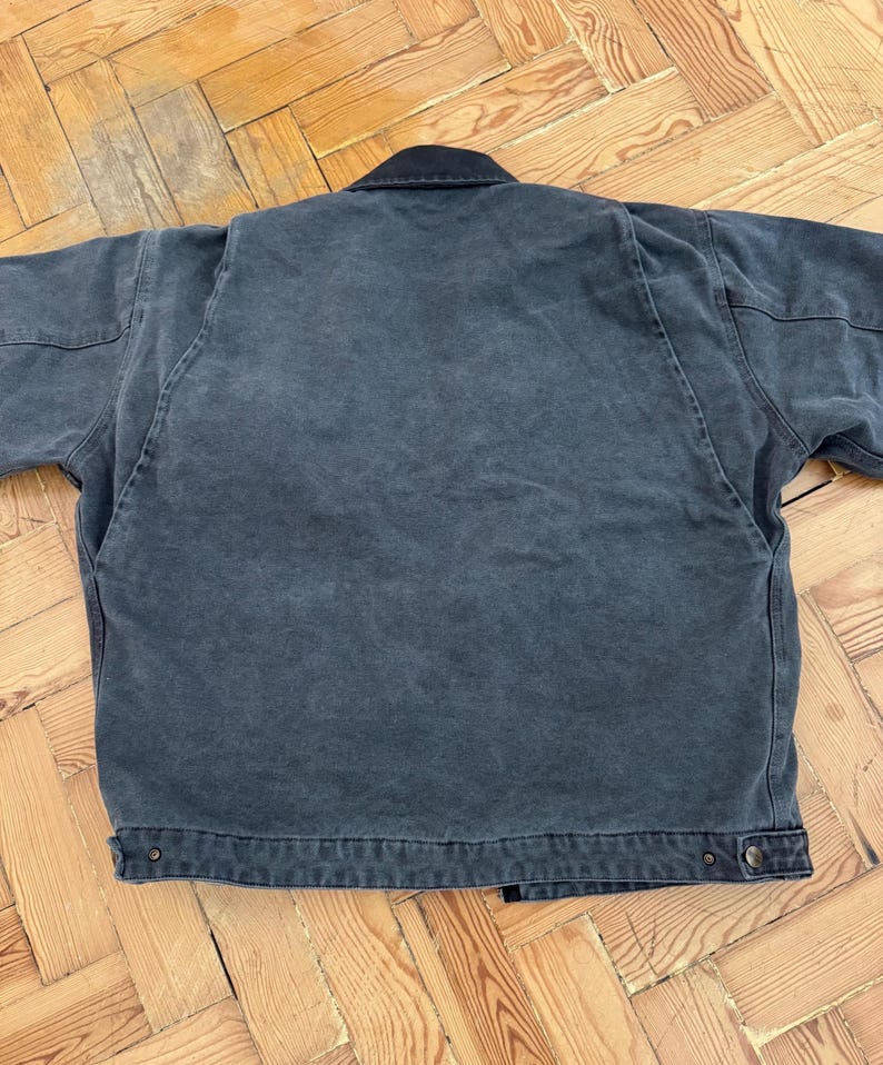 May include: A dark gray work jacket with a black collar. The jacket has a button closure and two front pockets. The fabric appears to be a durable cotton or canvas material, suitable for work or casual wear.