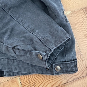 May include: Close-up of a dark gray work jacket. The jacket features a buttoned cuff and two metal buttons. The fabric has a textured appearance, and the stitching is visible along the seams. The jacket is lying on a wooden surface.