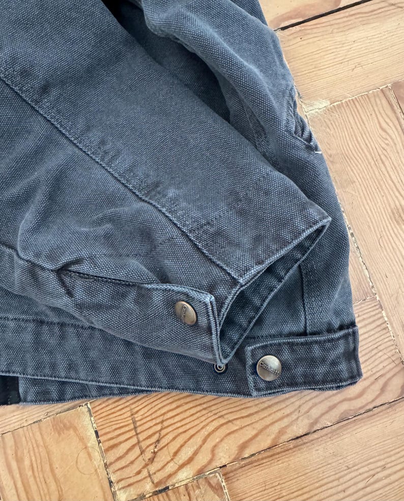 May include: Close-up of a dark gray work jacket. The jacket features a buttoned cuff and two metal buttons. The fabric has a textured appearance, and the stitching is visible along the seams. The jacket is lying on a wooden surface.