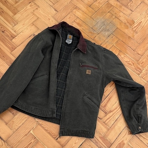 May include: A dark green jacket with a burgundy collar and a plaid interior. The jacket has a zipper closure, two front pockets, and a small square patch on the chest. The jacket is made of a durable material.