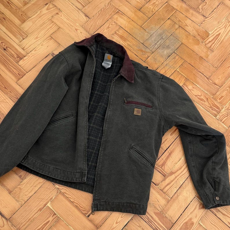 May include: A dark green work jacket with a corduroy collar and a Carhartt logo patch. The jacket has a zipper closure and multiple pockets.
