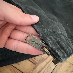 May include: Close-up of a zipper pull on a dark green jacket. The zipper pull is metal and has the word "Carhartt" engraved on it.