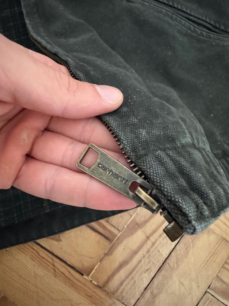 May include: Close-up of a zipper pull on a dark green jacket. The zipper pull is metal and has the word "Carhartt" engraved on it.