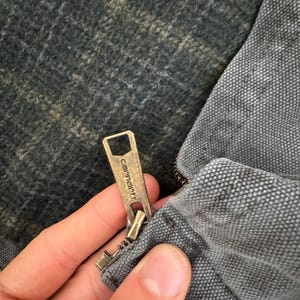 May include: Close-up of a Carhartt jacket zipper. The zipper is brass-colored and features the Carhartt logo. The jacket is gray and has a textured fabric. The interior lining is a dark, patterned material.