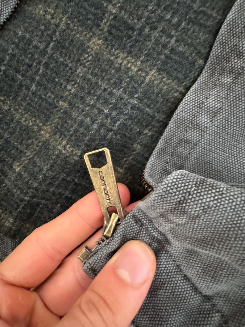 May include: Close-up of a Carhartt jacket zipper. The zipper is brass-colored and features the Carhartt logo. The jacket is gray and has a textured fabric. The interior lining is a dark, patterned material.