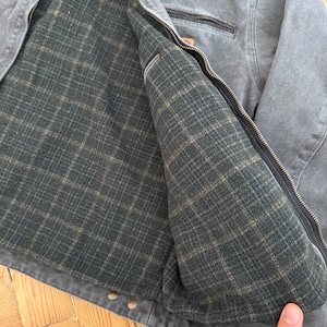 May include: Gray work jacket with a full zipper closure. The jacket features a dark green plaid lining and a small pocket on the chest. The jacket is made of a durable, heavy-duty material.