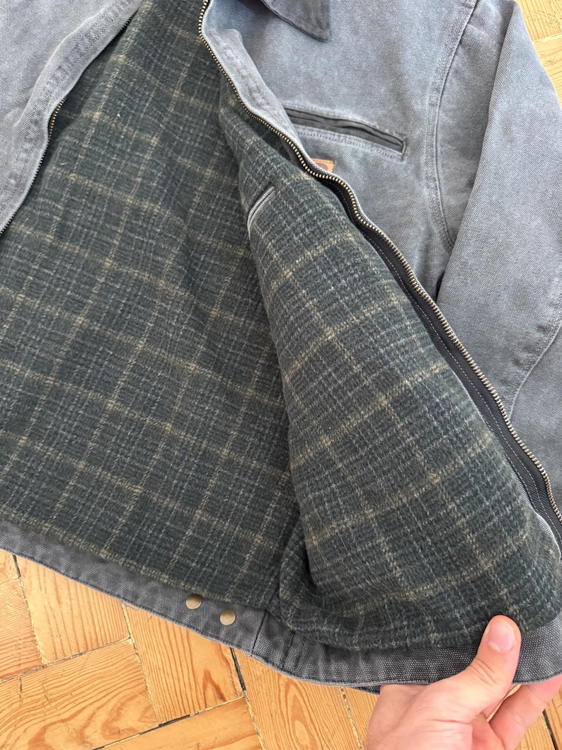 May include: Gray work jacket with a full zipper closure. The jacket features a dark green plaid lining and a small pocket on the chest. The jacket is made of a durable, heavy-duty material.