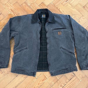 May include: A charcoal-colored work jacket with a black collar and a plaid interior. The jacket has two front pockets, a chest pocket with a zipper, and a small square logo patch. The jacket is laid flat on a wooden surface.