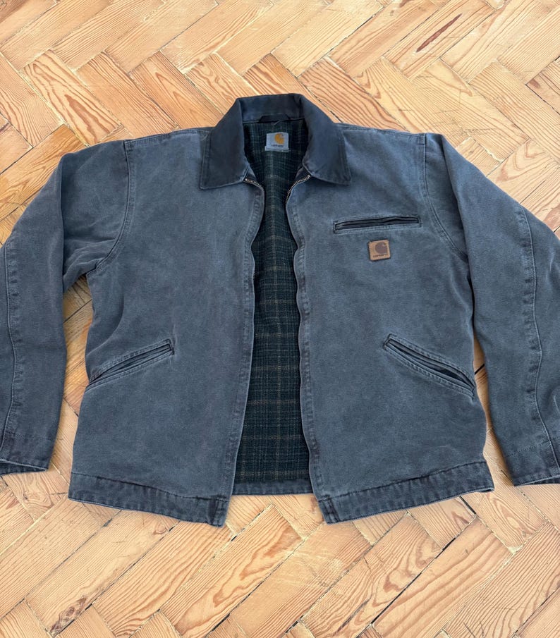 May include: A charcoal-colored work jacket with a black collar and a plaid interior. The jacket has two front pockets, a chest pocket with a zipper, and a small square logo patch. The jacket is laid flat on a wooden surface.
