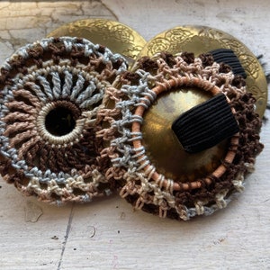 May include: Two brass cymbals with crocheted covers in brown, beige, and light blue. The cymbals have a decorative design and are attached together with a black elastic band.