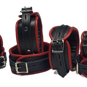 Può includere: Un set di manette in pelle nera con finiture rosse e fibbie argentate. Il set include manette, cavigliere e un collare. La pelle appare strutturata e le fibbie sono lucide.