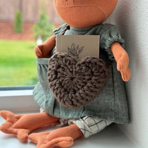 May include: A handmade doll with orange fabric arms and legs, wearing a green dress and plaid shorts. A brown crocheted heart-shaped purse is attached to the doll. The doll holds a small book with a leaf design.