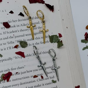 May include: Two pairs of dagger-shaped earrings. One pair is gold-colored, the other is silver-colored. Both pairs have a hoop closure. The earrings are displayed on an open book with scattered rose petals.
