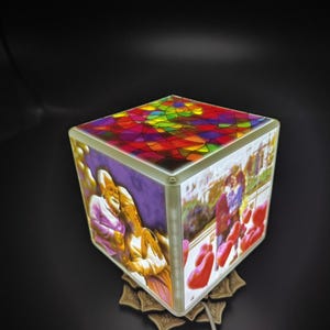 Può includere: Una lampada a forma di cubo con un piano in vetro colorato e immagini sui lati. I lati mostrano fotografie di persone. La lampada poggia su una base decorativa a forma di fiore. La parte superiore della lampada presenta un design colorato in vetro colorato.