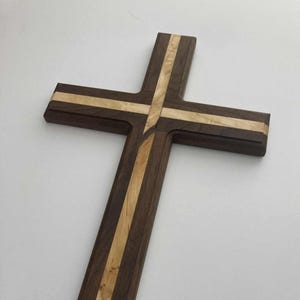 Może przedstawiać: Drewniany krzyż z ciemnobrązową podstawą i jasnobrązowym paskiem pośrodku. Krzyż wykonany jest z dwóch połączonych ze sobą kawałków drewna. Krzyż ma około 30 cm długości i 20 cm szerokości.