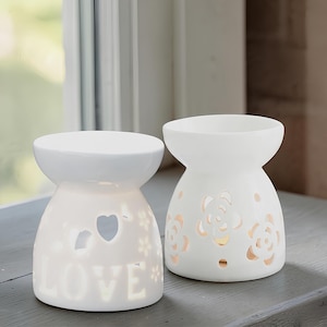 Könnte beinhalten: Zwei weiße Keramik-Duftlampen. Eine Lampe hat ein Herz und das Wort "LOVE" ausgeschnitten. Die andere hat Blumenausschnitte. Beide haben eine runde Oberseite und einen breiteren Sockel. Sie stehen auf einer grauen Oberfläche in der Nähe eines Fensters.