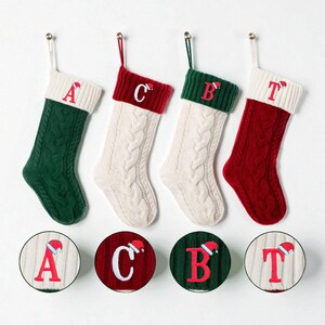 Op de afbeelding: Vier kabelgebreide kerstsokken in groen, crème en rood, elk met een andere letter in het rood geborduurd. De sokken hangen aan gouden haken. Bijpassende ronde ornamenten met letters en kerstmutsen eronder.