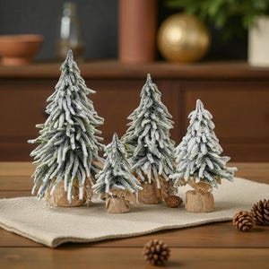 Può includere: Una collezione di cinque alberi di Natale artificiali in miniatura con effetto neve. Gli alberi sono verdi con accenti bianchi e sono inseriti in basi avvolte in juta. La composizione è su un runner di lino color neutro, con pigne sparse.