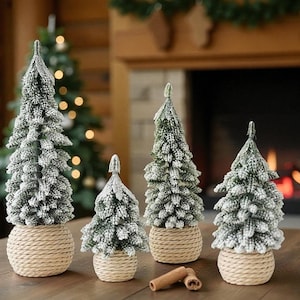 Puede incluir: Cuatro árboles de Navidad decorativos en miniatura con apariencia nevada. Cada árbol es verde con detalles blancos y se asienta sobre una base tejida de color beige. Una ramita de canela descansa sobre la superficie de madera frente a los árboles.