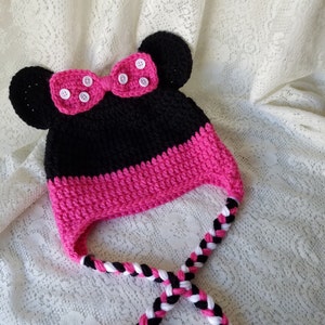Könnte beinhalten: Eine rosa und schwarze Häkelmütze mit Schleife und Mäuseohren. Die Mütze hat einen langen geflochtenen Riemen.