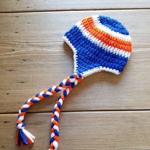 May include: A blue, white, and orange crocheted hat with a long braided cord. The hat has a pointed top and a flap that covers the ears.