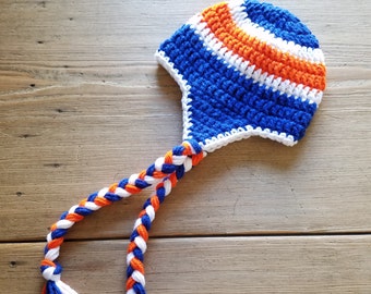 May include: A blue, white, and orange crocheted hat with a long braided cord. The hat has a pointed top and a flap that covers the ears.