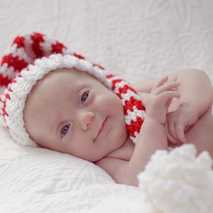 Puede incluir: Un bebé con un gorro de Papá Noel de crochet rojo y blanco y una manta blanca. El bebé está acostado sobre una manta blanca y tiene ojos azules.