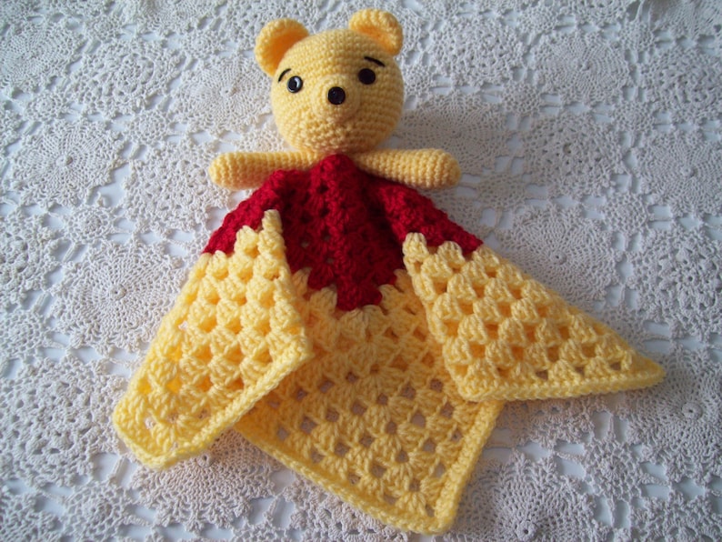 May include: A crocheted baby blanket with a yellow and red pattern. A yellow crocheted bear is attached to the blanket.