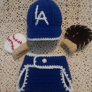 May include: A crocheted baby set in blue, white, and brown, featuring a baseball cap with "LA" in white, a baseball-themed mitten, a baseball, and a diaper cover with a baseball button. The set is handmade.