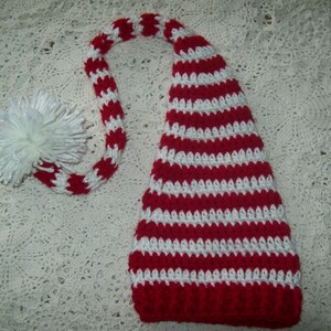 Puede incluir: Un gorro de elfo de crochet a rayas rojas y blancas con un pompón blanco en el extremo de la cola larga.
